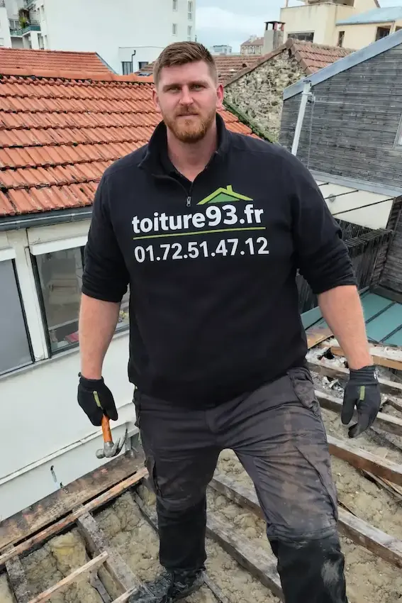 Un couvreur barbu portant un pull noir avec le logo "toiture93.fr" se tient debout sur une structure de toit en cours de rénovation. Il tient un marteau et porte des gants de protection. En arrière-plan, on aperçoit des toits en tuiles et des bâtiments urbains.